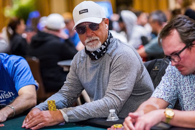 A player in a white cap, sunglasses and a grey long-sleeve top sits at a tournament table with his hands on the felt beside a stack of chips, other players around him