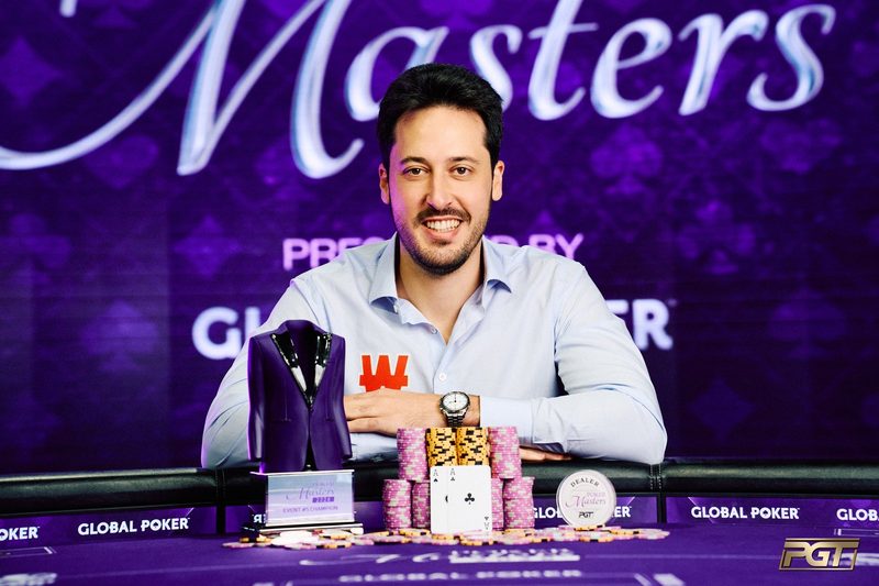 Adrian Mateos at the Poker Masters winner's table, smiling behind stacks of chips and the purple jacket trophy