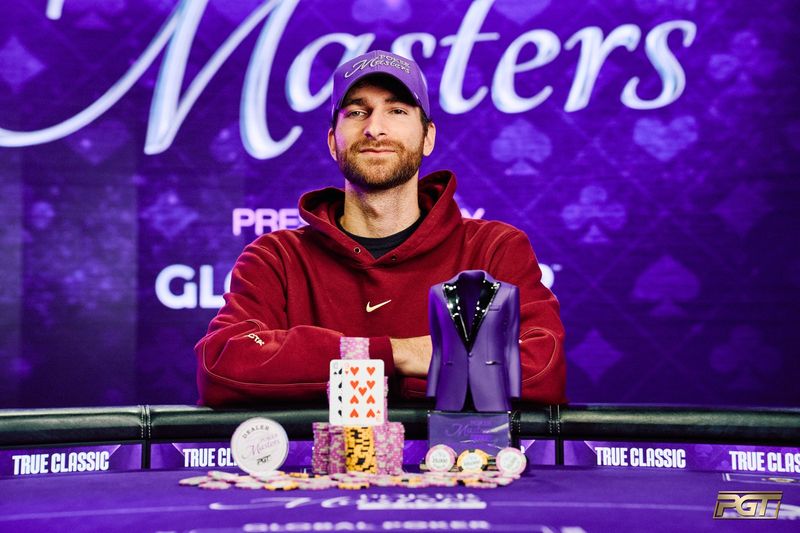 Justin Vaysman in a purple Poker Masters cap and a dark red hoodie at the winner's table, arms folded behind stacks of chips, the ten and nine of hearts and the purple jacket trophy