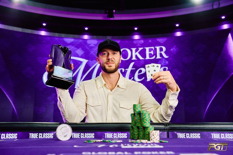 Sean Winter in a black cap and cream shirt holding up the purple jacket trophy and the six-five of spades and clubs behind a stack of chips in front of a Poker Masters backdrop